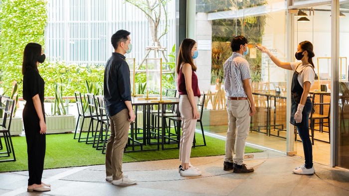 Asian waitress wear protective face mask scan customer temperature by thermometer before entering the restaurant to protect infection from coronavirus covid-19, social distancing concept