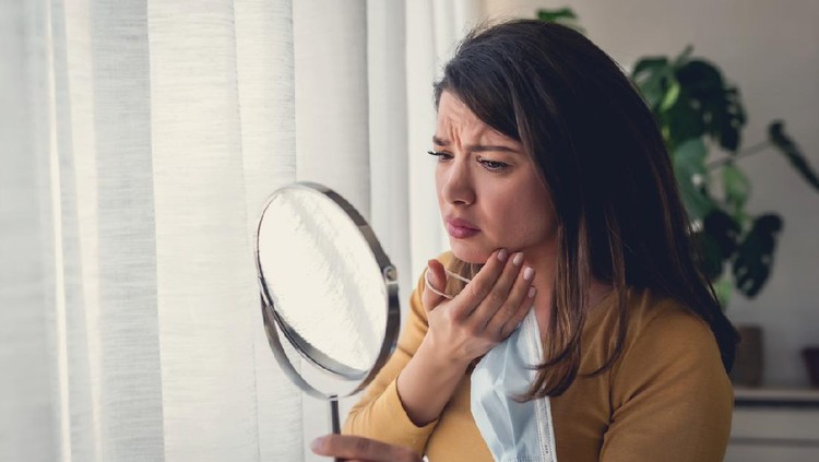 Ilustrasi Bekas Jerawat Woman looking herself in the mirror at home standing by the big window. She is concerned about acne, maskne