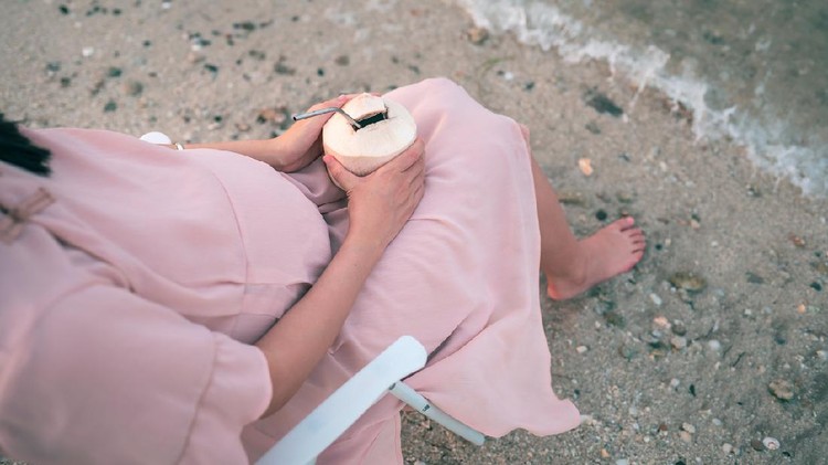 Ilustrasi ibu hamil minum air kelapa Cheerful pregnant woman having fresh coconut on the beach sitting on a picnic foldable chair by the seaside and enjoying the sunset. Summer vacation during pregnancy and outdoors lifestyle