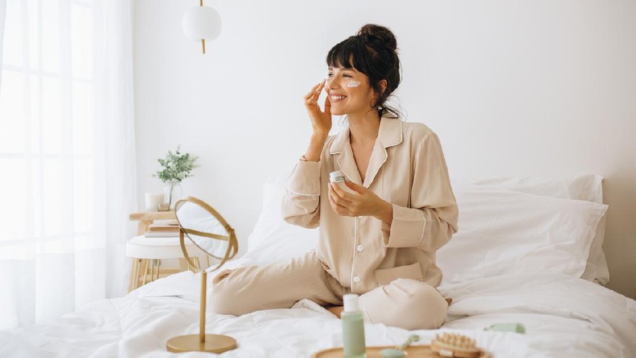 Happy woman doing routine skin care at home with beauty products. Woman sitting on bed at home and applying face cream.