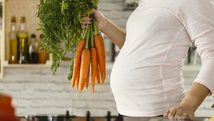 Pregnant mid adult woman holding bunch of carrots in the kitchen, preparing food.
