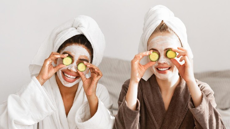 Playful multiracial girlfriends in bathrobes and face masks on covering eyes with avocado halves, beauty and spa concept, panorama