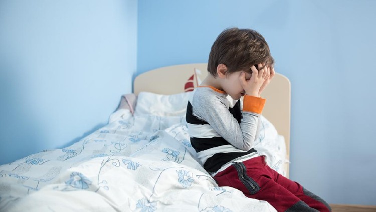 Sad, upset little boy sitting on the edge of his bed. Covering his face with hands.