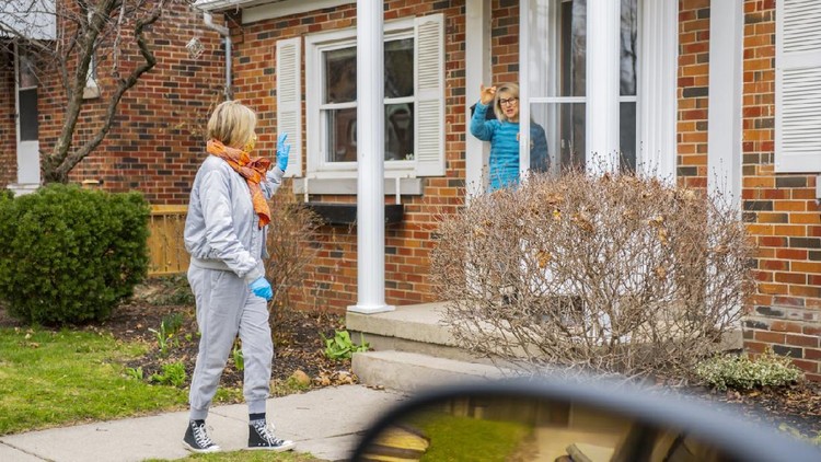 Two women waving good bye on the front lawn of a residential home.  The woman leaving is wearing protective gloves and a face mask.  They are practicing social distancing.