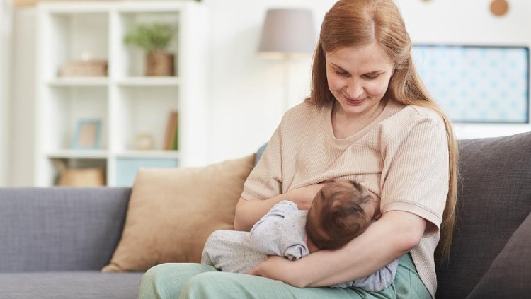 menyusui Breastfeeding baby near crip. Pretty mother holding her newborn child. Mom nursing baby. Beautiful woman and new born relax at home in nursery interior. Blond mother breast feeding baby.