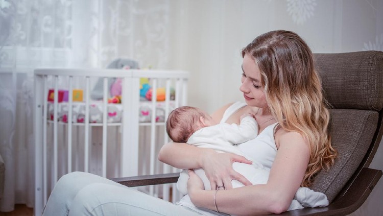 Breastfeeding baby near crip. Pretty mother holding her newborn child. Mom nursing baby. Beautiful woman and new born relax at home in nursery interior. Blond mother breast feeding baby.
