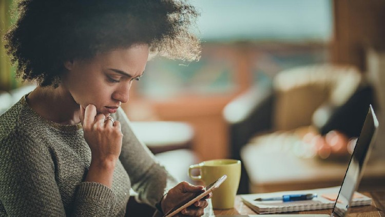 Young black woman reading a problematic text message on her mobile phone while working at home.