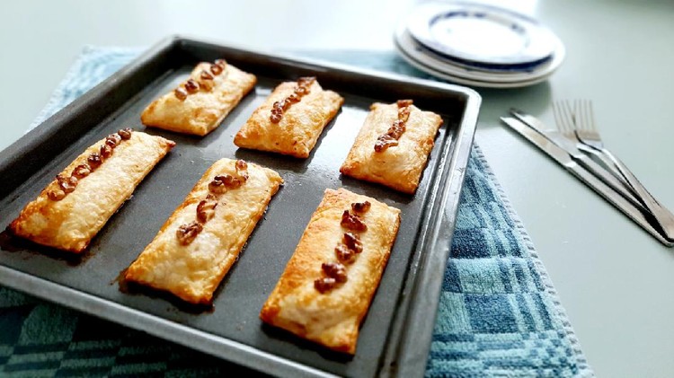 Baked puff pastry homemade stuffed with goat cheese, honey and walnuts. 6 pieces on a baking sheet fresh from the oven. Group of objects. Golden brown on grey. Table blue green with forks and knives and dome plates. Focus on foreground. Daylight.