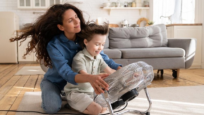 Cheerful young mom or nanny have fun with preschool boy at home sitting together in front of big fan dlowing cooling wind in living room. Carefree caring woman of 30 spend time with little son bonding