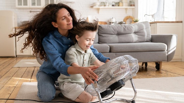 Cheerful young mom or nanny have fun with preschool boy at home sitting together in front of big fan dlowing cooling wind in living room. Carefree caring woman of 30 spend time with little son bonding