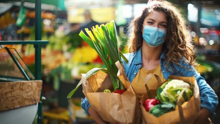 Closeup side view of a late 20's woman going grocery shopping during coronavirus pandemic. She's at a farmer's market  with bags full of fresh fruit and vegetables, smiling and looking at the camera,