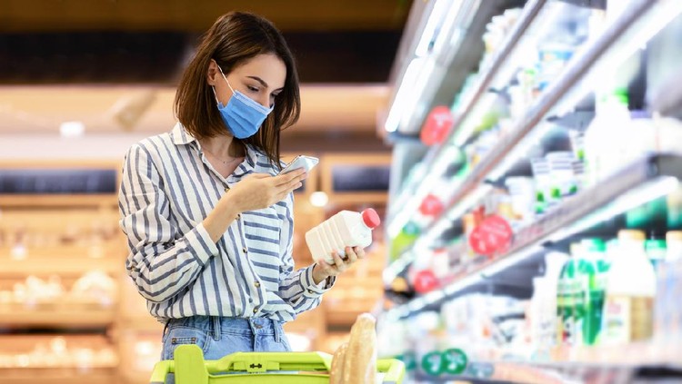Young woman in disposable face mask taking dairy products from shelf in the supermarket, holding bottle and smartphone, scanning bar code on product through mobile phone, walking with trolley cart
