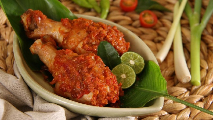 Ayam Rica-Rica, the popular and traditional dish from Manado, chicken braised in spicy red chili paste. Two drumsticks are arranged on a small ceramic plate that has been lined with a banana leaf. Two lime halves and a lime leaf are used to garnish the dish. The plate is placed on a woven bamboo place mat. Some of the herbs and spices are also placed on the place mat.