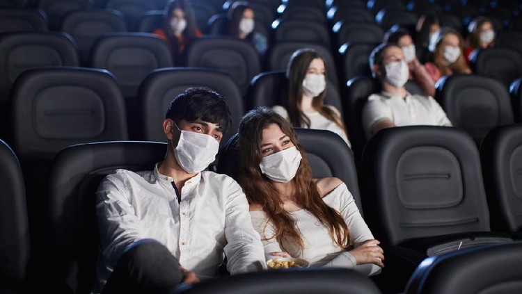 Selective focus of man and woman enjoying film during world pandemic, looking at screen. Young caucasian audience watching movie in cinema, wearing white face masks. Social distancing concept.