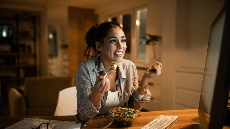 Smiling woman reading an e-mail on desktop PC and eating salad in the evening at home.