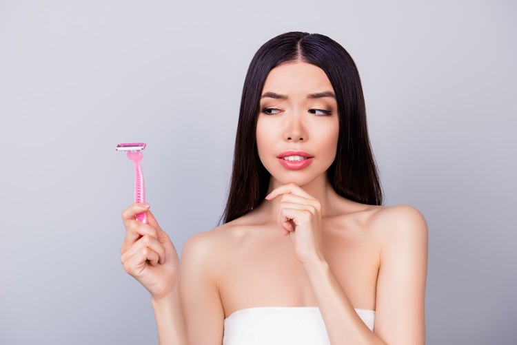 Young unsure nervous asian girl with pink shaver is standing isolated on a grey background, wrapped in a white towel, doubting if she should use it