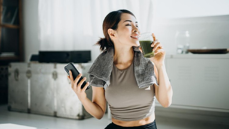 Young Asian sports woman resting and having green smoothie after exercising at home in the morning, using smartphone