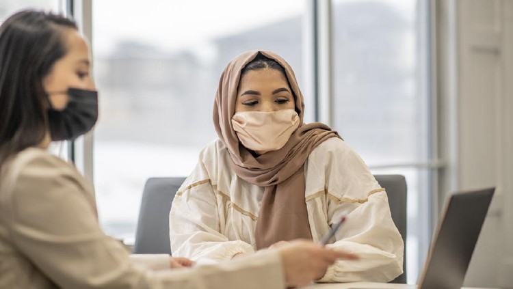 A Muslim woman is sitting at her doctor's office during an appointment. Her doctor is pointing to her laptop's screen to explain the test results to her patient. They are both wearing a face mask during the coronavirus pandemic to keep each other safe.