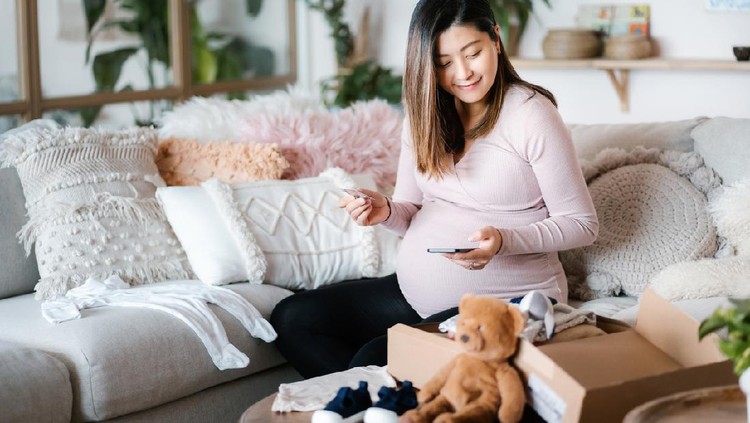 Smiling Asian pregnant woman shopping online with smartphone and making mobile payment with credit card in the living room. A delivery package of baby clothing and toys on coffee table. Time to get some baby essentials for her unborn baby!