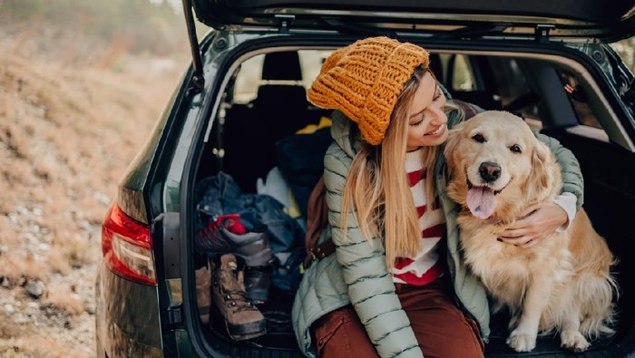 Photo of a young smiling woman and her dog sitting the trunk of a car on a beautiful autumn day; taking a short break during their road trip.