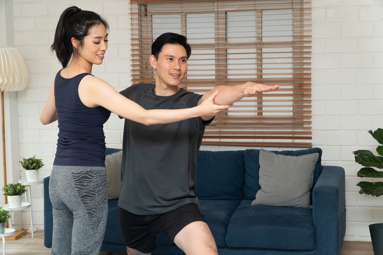 Healthy young Asian couple exercises in home and wife teaching her husband for exercise in COVID-19 quarantine at home. A new normal lifestyle and healthy exercise can be done anywhere.