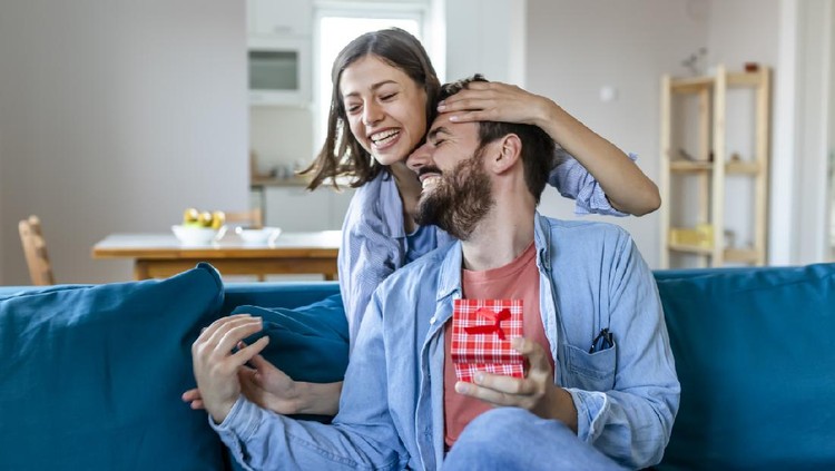 Beautiful Girl Surprise Her Boyfriend With Gift Celebrating Anniversary While He Is Sitting on the Sofa in the Living Room. Romantic Surprise for Husband Boyfriend, Happy Birthday Concept