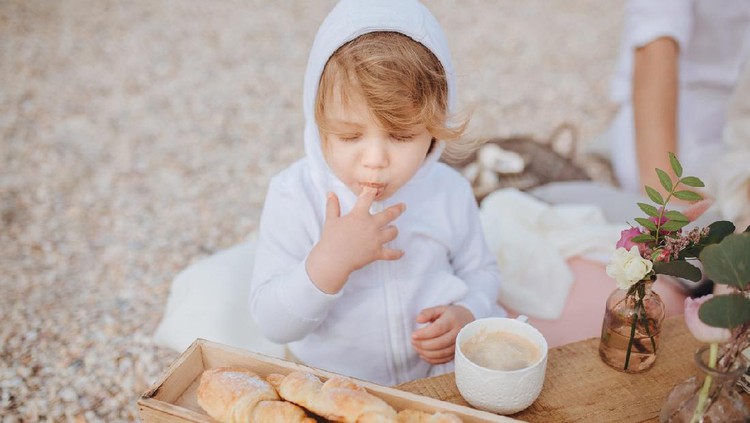 ilustrasi anak Child licks his finger with closed eyes of pleasure standing at the table with croissants