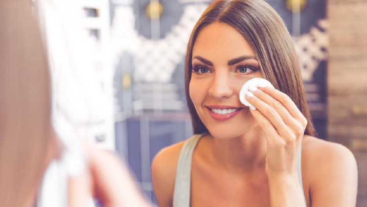 Beautiful young woman is cleaning her face using a cotton disc and smiling while looking in the mirror in the bathroom