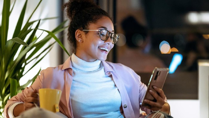 Shot of happiness young entrepreneur woman using her smart phone while drinking a cup of coffee sitting on couch in the office.
