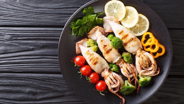 grilled calamari with tentacles with tomatoes, broccoli, lemon and pepper closeup on a plate on a table. Horizontal top view from above