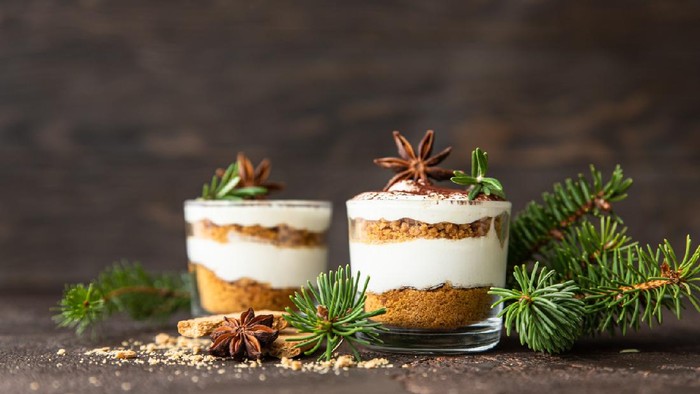 Layered dessert in glass jar with cookie crumble and whipped cream decorated with rosemary and anise, dark brown background. No bake cheesecake, trifle or pudding.