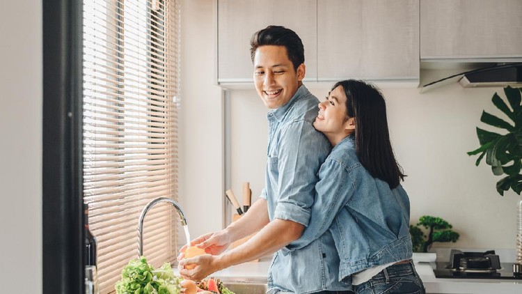 Asian couple lovers hug in the kitchen while cooking at home. A woman hugs a man who is washing vegetables from the back.