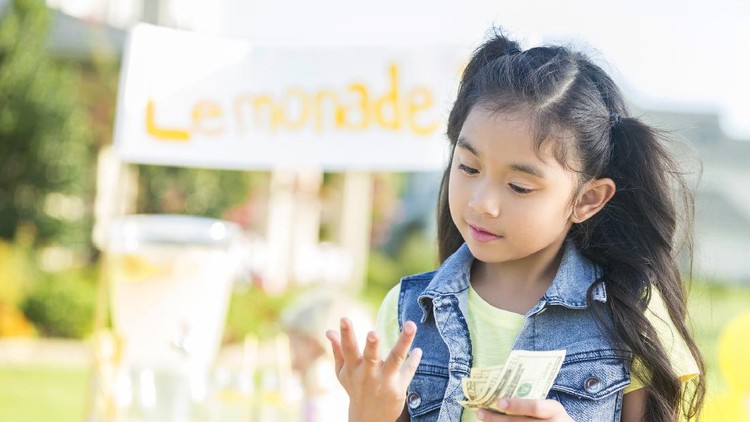 Pretty girl counts on her fingers while holding cash from her lemonade sales. The lemonade stand is in the background.