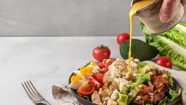 A hand pouring salad dressing into cobb salad with chicken, avocado, bacon, blue cheese, tomato and eggs. Keto diet food. Classic American dish