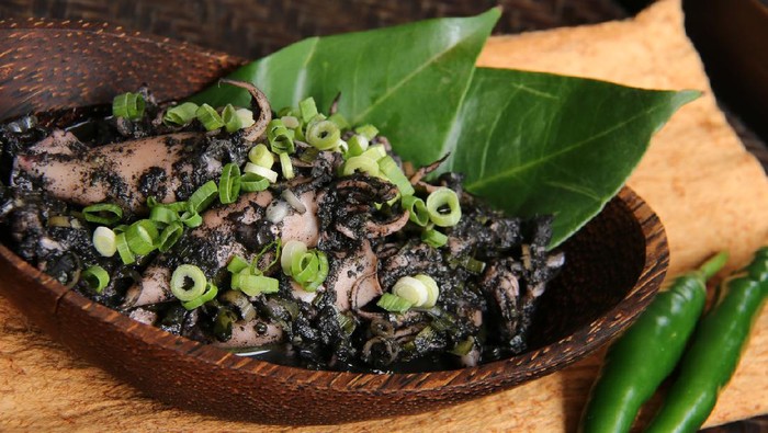 Cumi Woku Tinta, a popular Minahasan seafood dish of squids braised with woku spices and squid ink from Manado, North Sulawesi. It is served in an oval wooden bowl made of arenga wood, lined with salam bay leaves and garnished with chopped scallion.