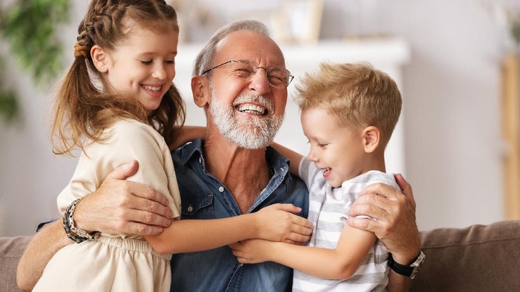 Cheerful aged man smiling and embracing cute boy and girl while resting on couch at home together