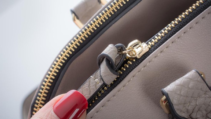 Closing a beige leather purse. Close up of a thumb with red nail pulling a golden zipper with a beige leather cord