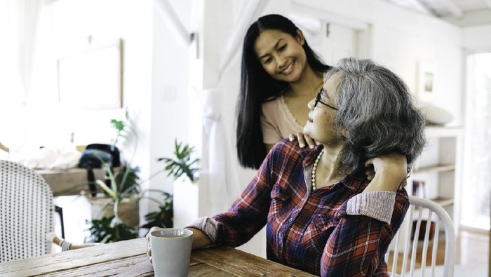 Beautiful adult granddaughter hugging beautiful senior grandmother while sitting on the chair drinking coffee