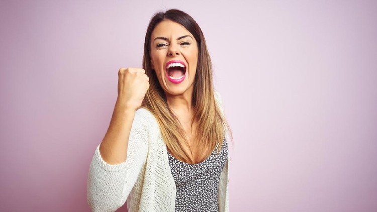 Young beautiful woman standing over pink isolated background angry and mad raising fist frustrated and furious while shouting with anger. Rage and aggressive concept.