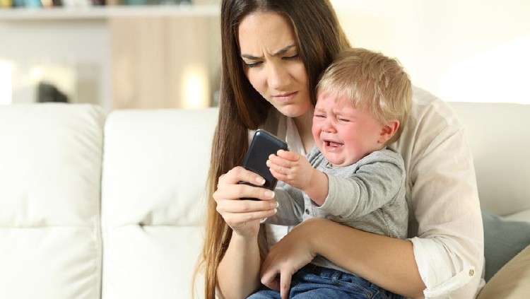 Baby having a tantrum and fighting with his mother for a smart phone sitting on a couch in the living room at home