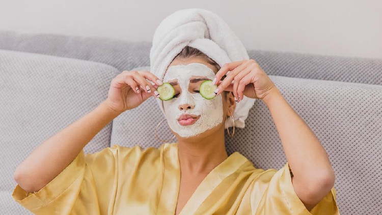 Beautiful young woman doing spa treatments at home, lying on the couch. Holds pieces of cucumbers in the arms above the eyes. Dressed in a bathrobe and a towel on the head.