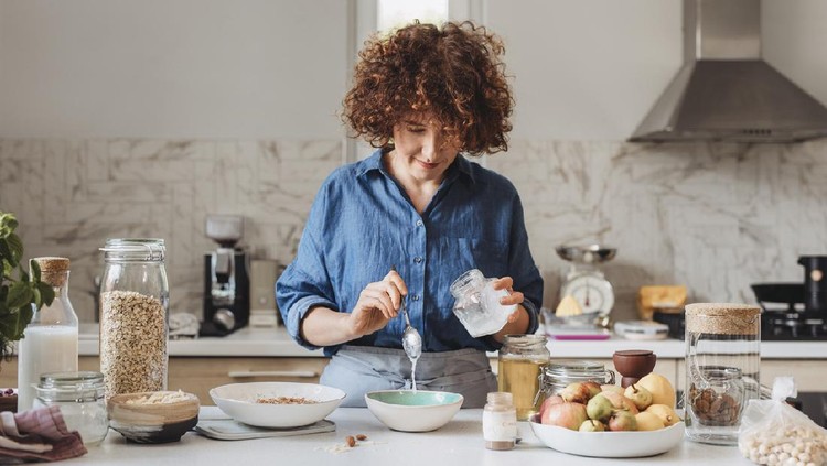 Homemade baked granola: woman adding ingredients into a bowl of rolled oats in the kitchen.