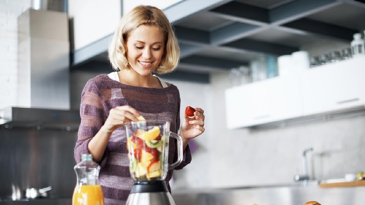 A young woman smiling happily while placing slices of fresh fruit into her blender