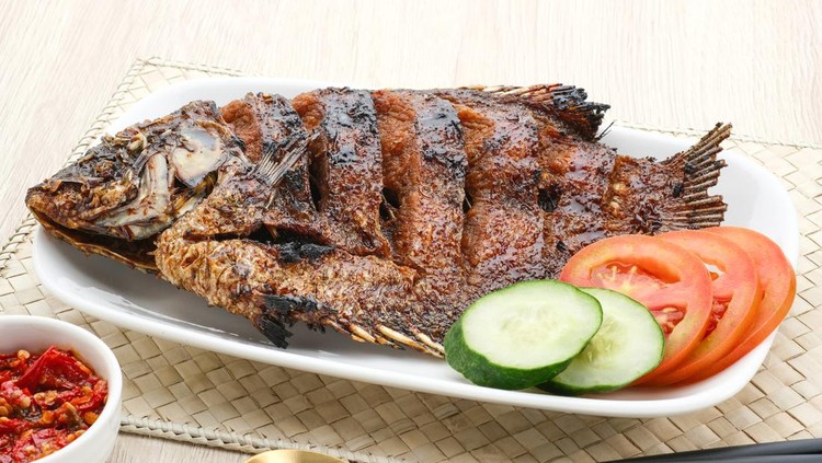 Gurame Bakar Madu, Grilled Gourami with honey and soy sauce. Served with sambal (chilli sauce), cucumber and tomatoes in white plate. Close up and selective focus image.