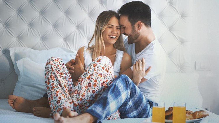 Closeup front view of late 20's couple relaxing in bed and having some juice and bagels. They are laughing and having some light conversation, it's weekend morning and they have all the day for themselves.