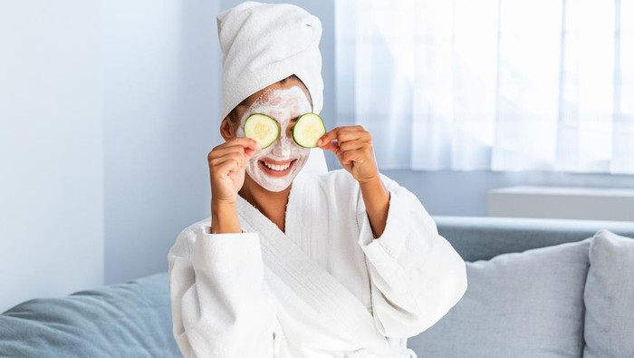 Woman with facial mask and cucumber slices in her hands. Beautiful young woman with facial mask on her face holding slices of fresh cucumber. Young woman with clay facial mask holding cucumber slices