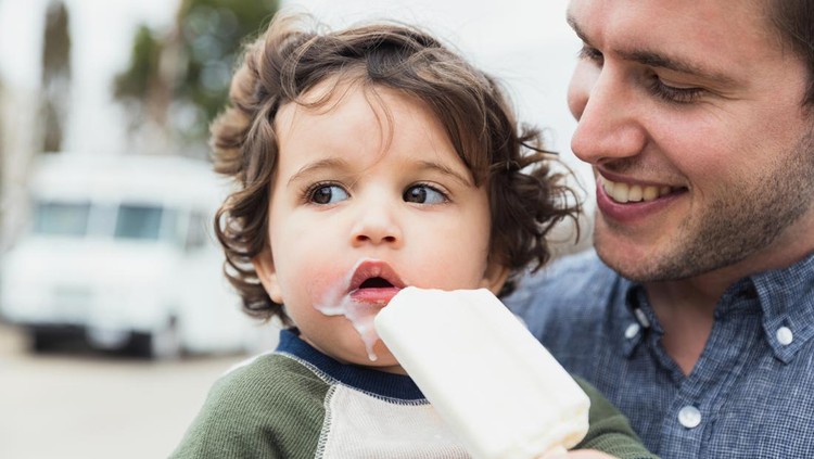 An adorable toddler boy with a messy mouth looks to the side while eating an ice cream treat. His dad smiles while holding the ice cream treat.