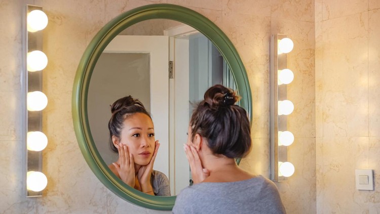 Rear view shot of a worried East Asian woman putting her hands on her cheek, checking her face skin from aging sign. She's standing in front a mirror with lights on both side.