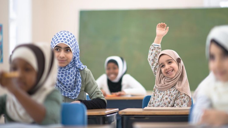 A young Muslim elementary student sits at her desk with her hand raised to ask the teacher a question.  She is smiling, wearing casual clothing and a head scarf.