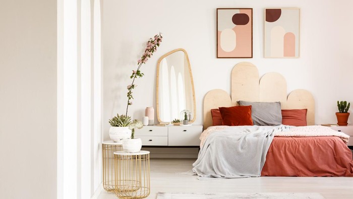 Real photo of a wide bed standing between a shelf with a plant and  a shelf with a mirror in a rusty bedroom interior with two posters on a wall and two gold, small tables with plants